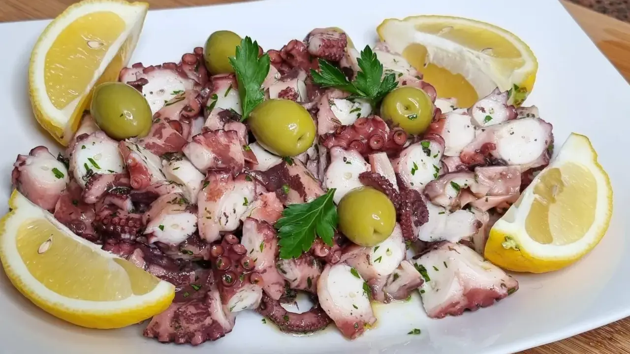 Octopus salad with olives, parsley, and lemon wedges on a white plate.