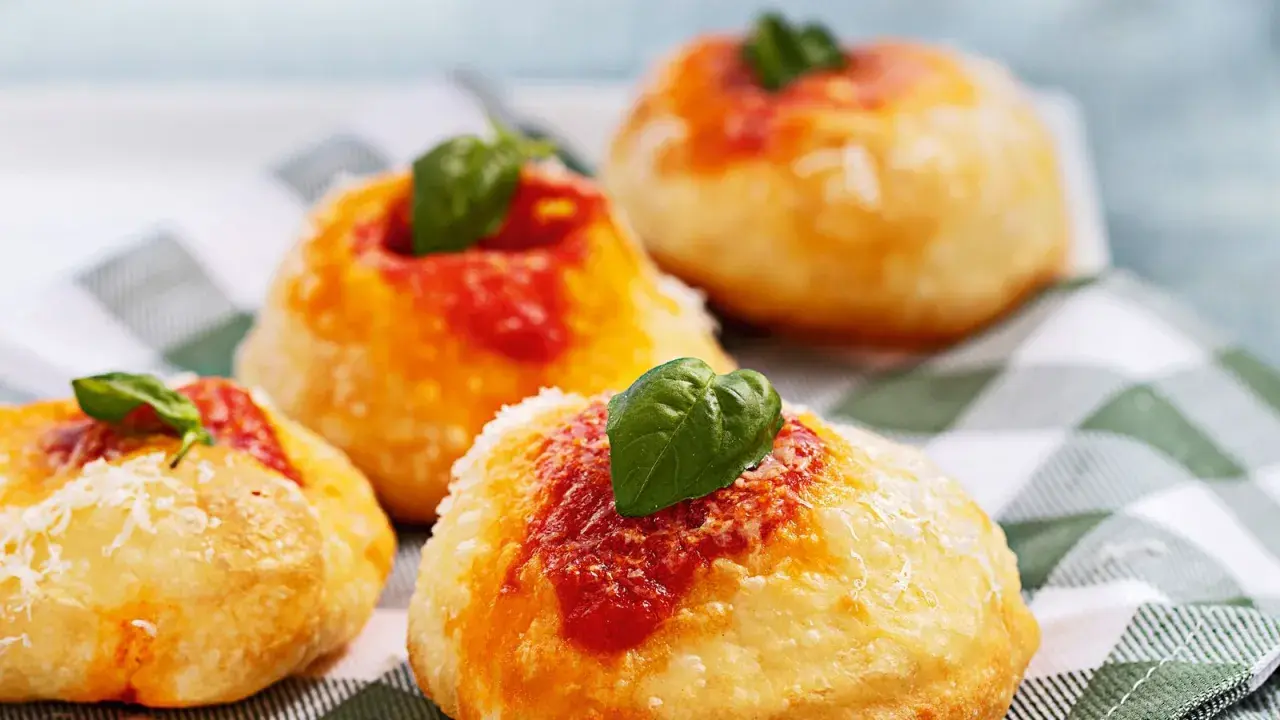 Four small, round Italian pastries topped with tomato sauce, grated cheese, and fresh basil leaves on a checkered cloth.