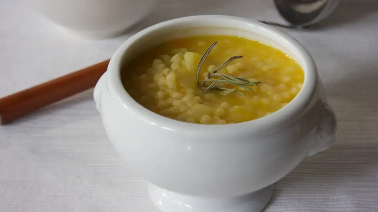 A white bowl of creamy potato soup with small pasta and a sprig of rosemary.