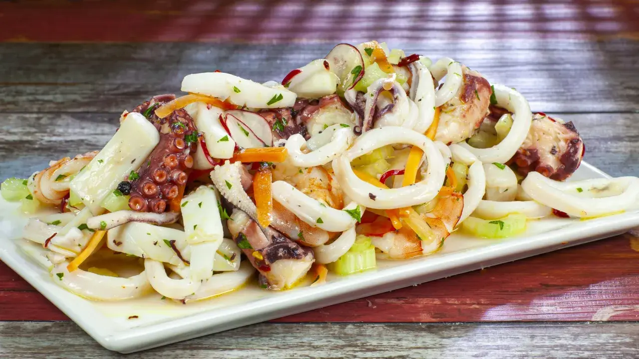 A white platter of seafood salad with octopus, calamari rings, shrimp, and vegetables on a wooden table.