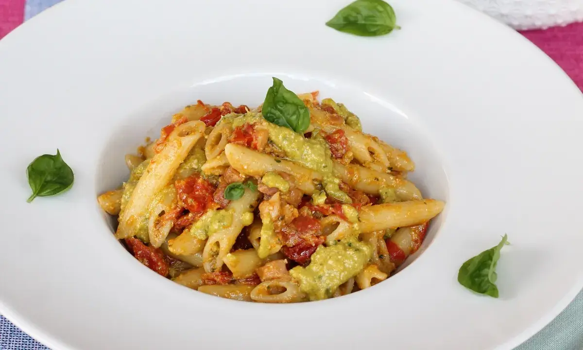 Penne pasta with a creamy green sauce, tomatoes, and basil leaves in a white bowl.