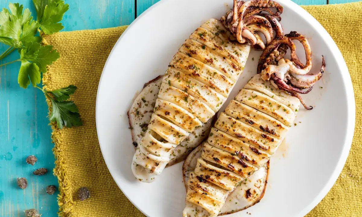 Two grilled calamari on a white plate, seasoned with herbs, on a blue wooden table.