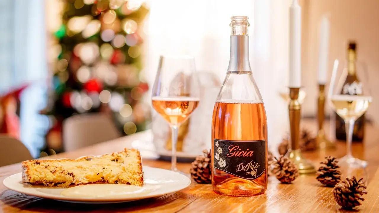 A slice of panettone with sauce, a bottle of rosé spumante, and pinecones on a wooden table.