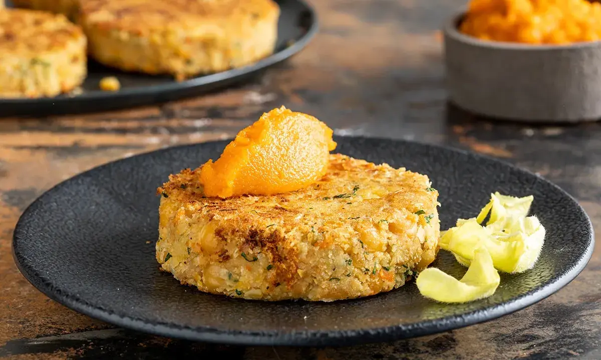 A golden-brown chickpea patty topped with orange puree on a black plate, with more patties in the background.