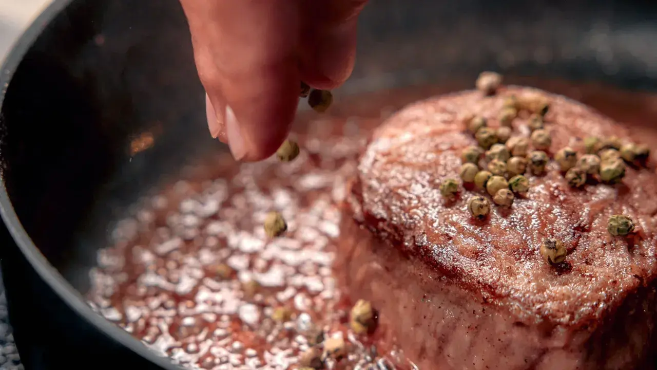 A hand sprinkles green peppercorns onto a seared steak in a pan.