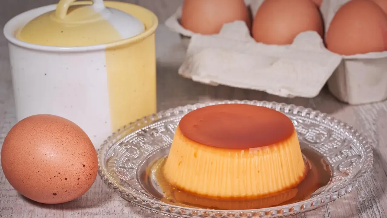 A crème caramel dessert on a decorative plate, with eggs and a ceramic jar in the background.