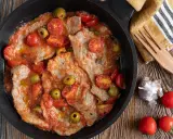 Thinly sliced meat in a tomato sauce with olives and cherry tomatoes in a black pan.