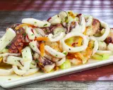 A white platter of seafood salad with octopus, calamari rings, shrimp, and vegetables on a wooden table.