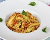Penne pasta with a creamy green sauce, tomatoes, and basil leaves in a white bowl.