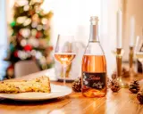 A slice of panettone with sauce, a bottle of rosé spumante, and pinecones on a wooden table.