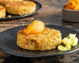 A golden-brown chickpea patty topped with orange puree on a black plate, with more patties in the background.