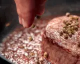 A hand sprinkles green peppercorns onto a seared steak in a pan.