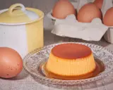 A crème caramel dessert on a decorative plate, with eggs and a ceramic jar in the background.
