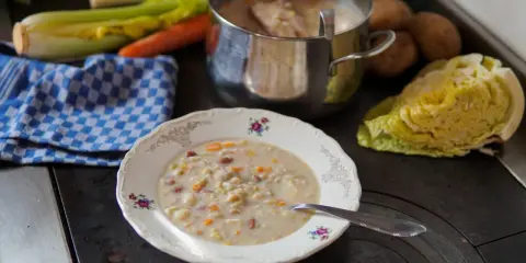 A bowl of hearty barley soup with vegetables and beans, next to a pot of soup and fresh ingredients.