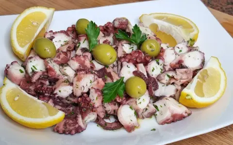 Octopus salad with olives, parsley, and lemon wedges on a white plate.