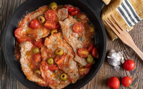 Thinly sliced meat in a tomato sauce with olives and cherry tomatoes in a black pan.