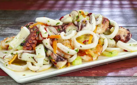 A white platter of seafood salad with octopus, calamari rings, shrimp, and vegetables on a wooden table.