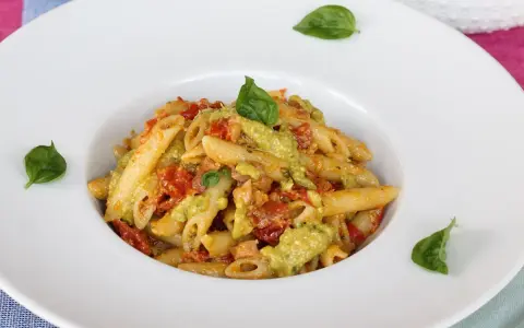 Penne pasta with a creamy green sauce, tomatoes, and basil leaves in a white bowl.