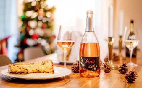 A slice of panettone with sauce, a bottle of rosé spumante, and pinecones on a wooden table.