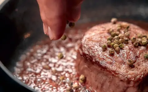 A hand sprinkles green peppercorns onto a seared steak in a pan.