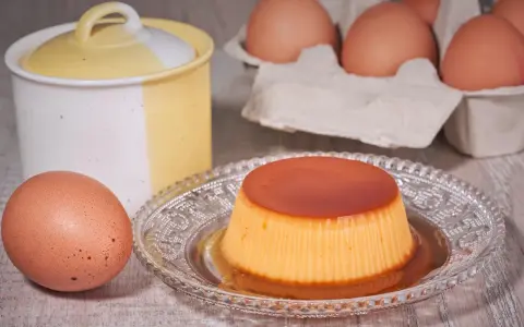 A crème caramel dessert on a decorative plate, with eggs and a ceramic jar in the background.