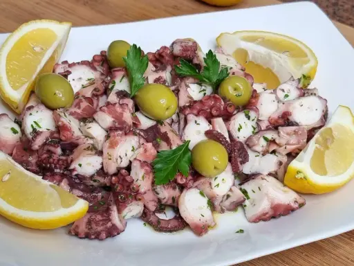 Octopus salad with olives, parsley, and lemon wedges on a white plate.