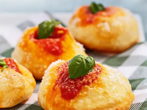Four small, round Italian pastries topped with tomato sauce, grated cheese, and fresh basil leaves on a checkered cloth.