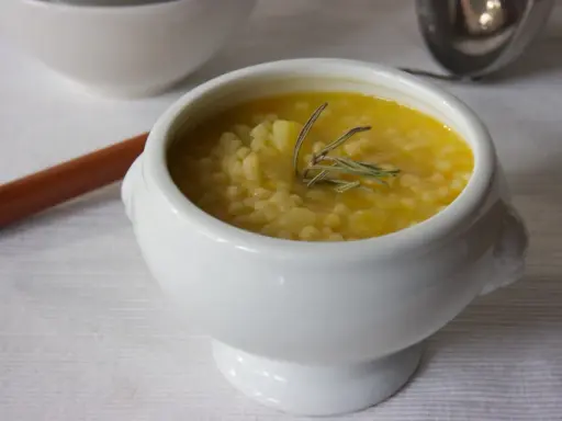 A white bowl of creamy potato soup with small pasta and a sprig of rosemary.