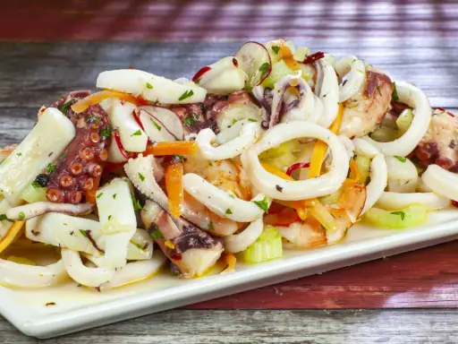 A white platter of seafood salad with octopus, calamari rings, shrimp, and vegetables on a wooden table.