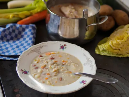 A bowl of hearty barley soup with vegetables and beans, next to a pot of soup and fresh ingredients.