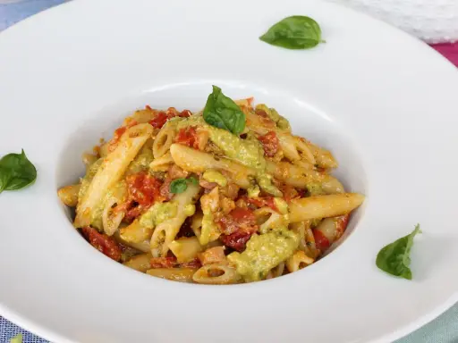 Penne pasta with a creamy green sauce, tomatoes, and basil leaves in a white bowl.