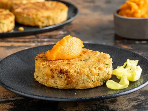 A golden-brown chickpea patty topped with orange puree on a black plate, with more patties in the background.