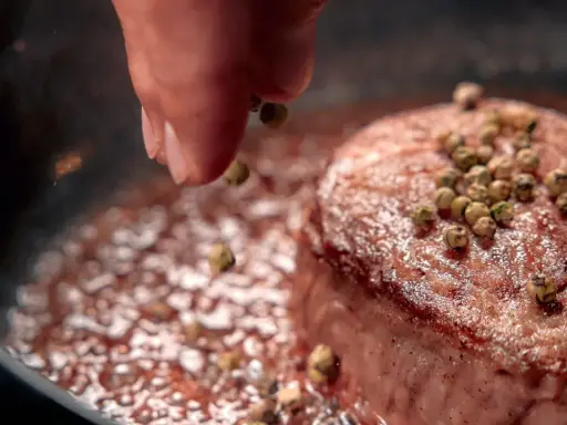 A hand sprinkles green peppercorns onto a seared steak in a pan.