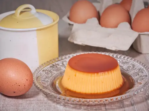 A crème caramel dessert on a decorative plate, with eggs and a ceramic jar in the background.
