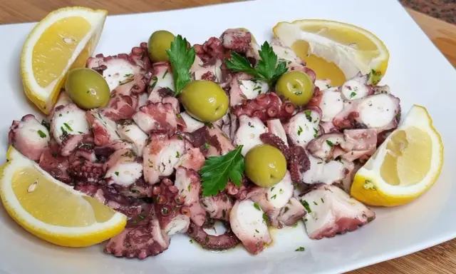 Octopus salad with olives, parsley, and lemon wedges on a white plate.