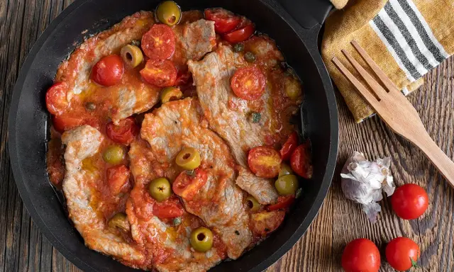 Thinly sliced meat in a tomato sauce with olives and cherry tomatoes in a black pan.