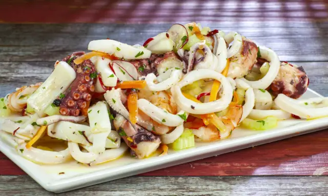 A white platter of seafood salad with octopus, calamari rings, shrimp, and vegetables on a wooden table.
