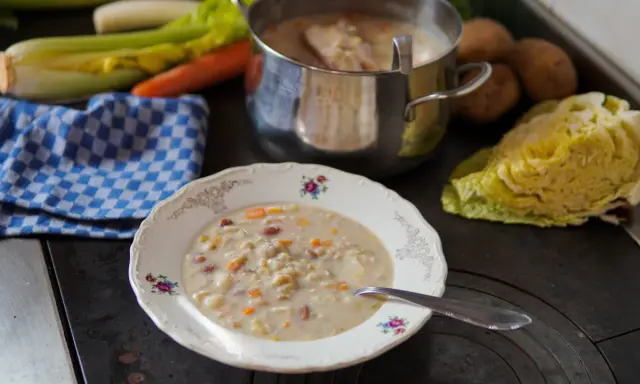 A bowl of hearty barley soup with vegetables and beans, next to a pot of soup and fresh ingredients.