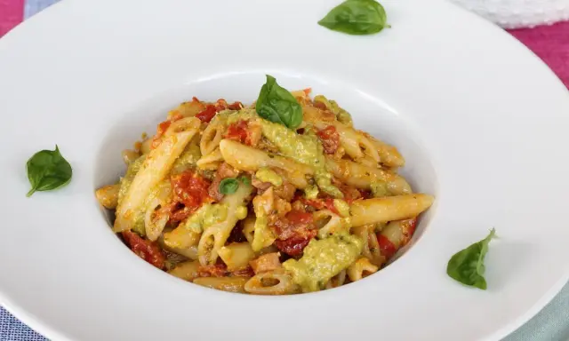 Penne pasta with a creamy green sauce, tomatoes, and basil leaves in a white bowl.