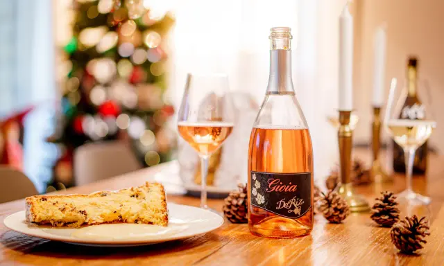 A slice of panettone with sauce, a bottle of rosé spumante, and pinecones on a wooden table.