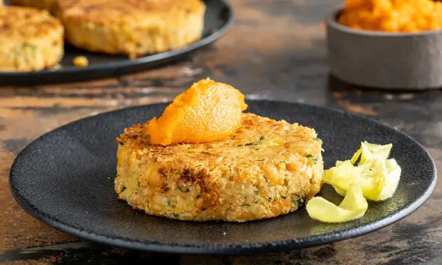 A golden-brown chickpea patty topped with orange puree on a black plate, with more patties in the background.