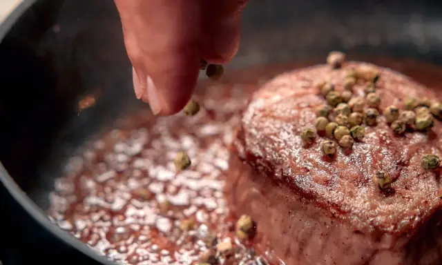 A hand sprinkles green peppercorns onto a seared steak in a pan.