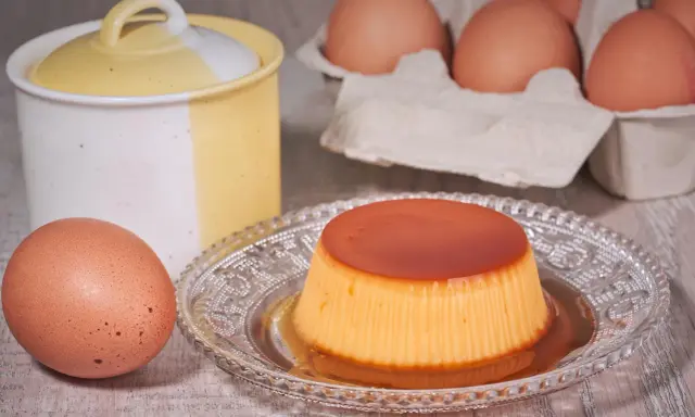 A crème caramel dessert on a decorative plate, with eggs and a ceramic jar in the background.