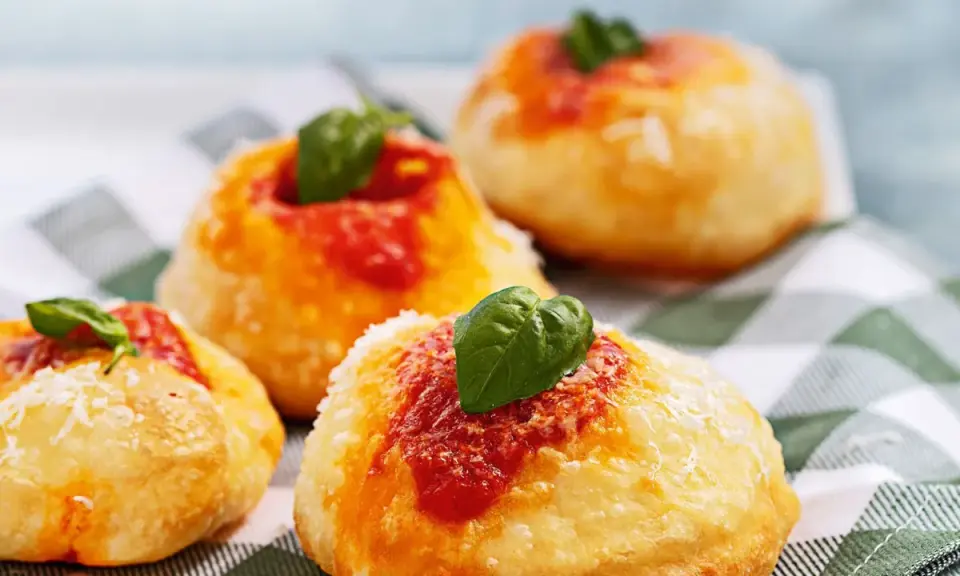 Four small, round Italian pastries topped with tomato sauce, grated cheese, and fresh basil leaves on a checkered cloth.