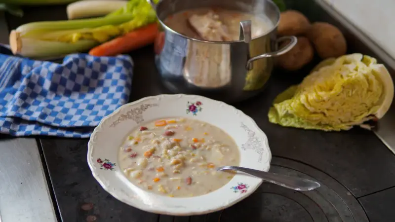 A bowl of hearty barley soup with vegetables and beans, next to a pot of soup and fresh ingredients.